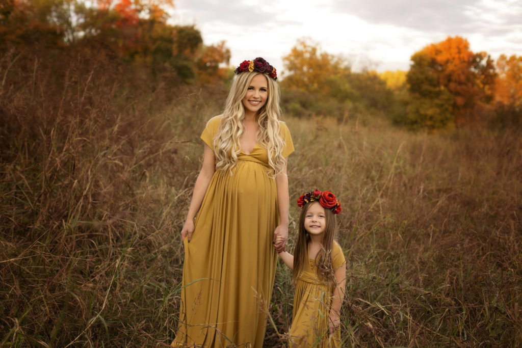 artistic maternity photos |Family Maternity Photo Shoot Ideas by popular Nashville lifestyle blog, Nashville Wifestyles: image of a pregnant woman holding her young daughter's hand as they stand in a field and both wear matching gold colored maxi dresses and floral crowns. 