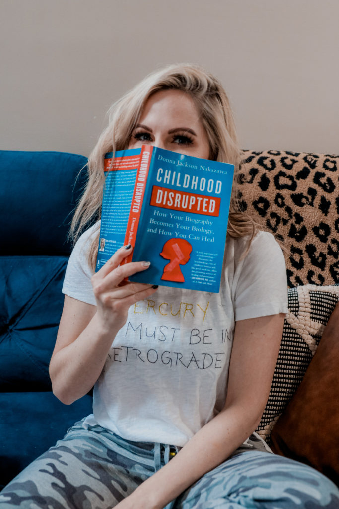 Self Healing Books by popular Nashville lifestyle blog, Nashville Wifestyles: image of a woman sitting on her blue couch and holding the Childhood Disrupted book. 