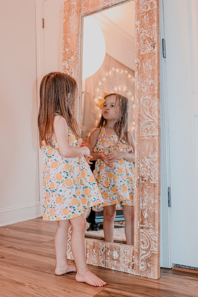 Toddler Room by popular Nashville motherhood blog, Nashville Wifestyles: image of a toddler age girl wearing a lemon print dress and looking at herself in a full body mirror. 