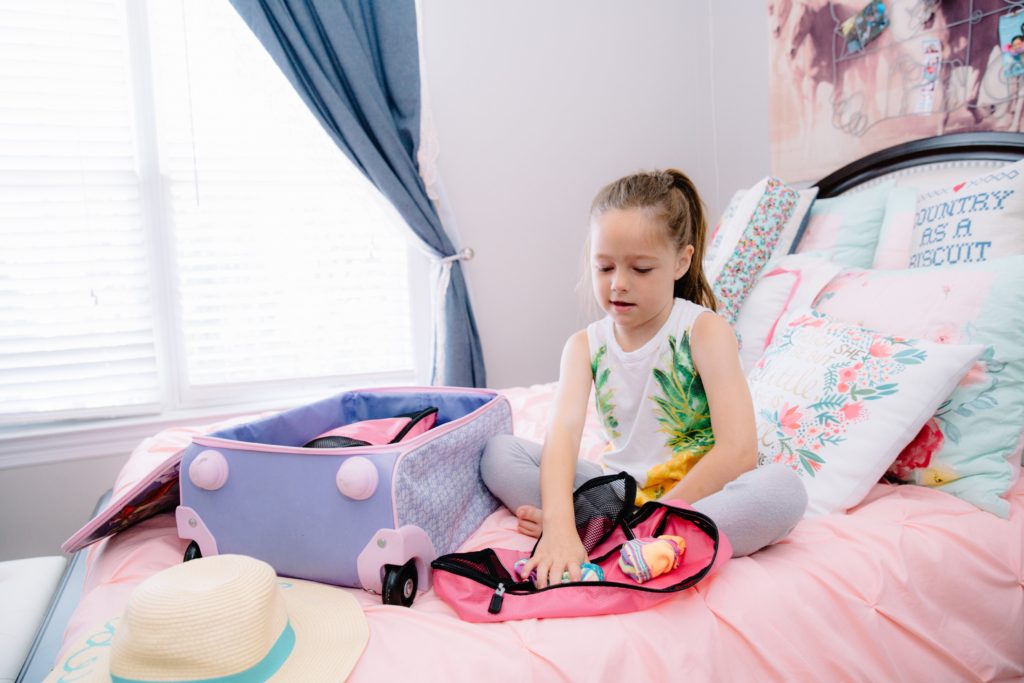 Travel Tips by popular Nashville travel blog, Nashville Wifestyles: image of a little girl sitting on her bed and packing her purple suitcase. 