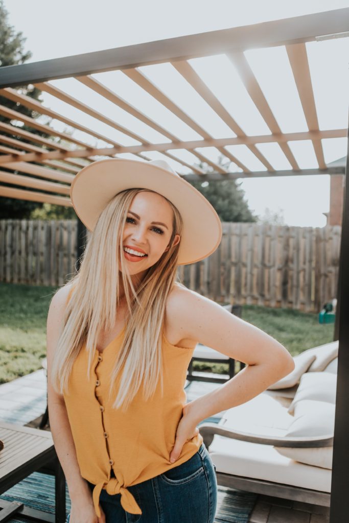 Sun Tanning Tips by popular Nashville beauty blog, Nashville Wifestyles: image of a woman standing outside under a pergola and wearing a pair of jeans, mustard yellow tank and tan felt fedora. 