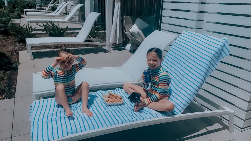 Soundwaves Water Park by popular Nashville blog, Nashville Wifestyles: image of two little girls wearing rainbow stripe rash guards and sitting on a white lounge chair while they eat chicken fingers and french fries. 