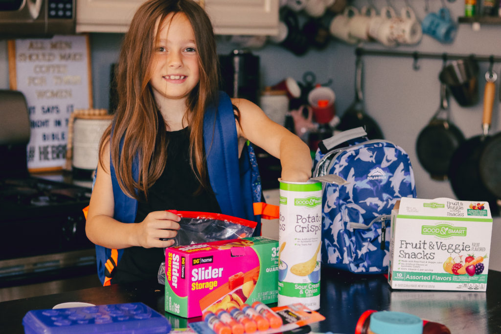Back to School Tips by popular Nashville lifestyle blog, Nashville Wifestyles: image of a girl opening a ziplock bag box while wearing a Dollar General backpack and standing next to a Dollar General lunch box. 