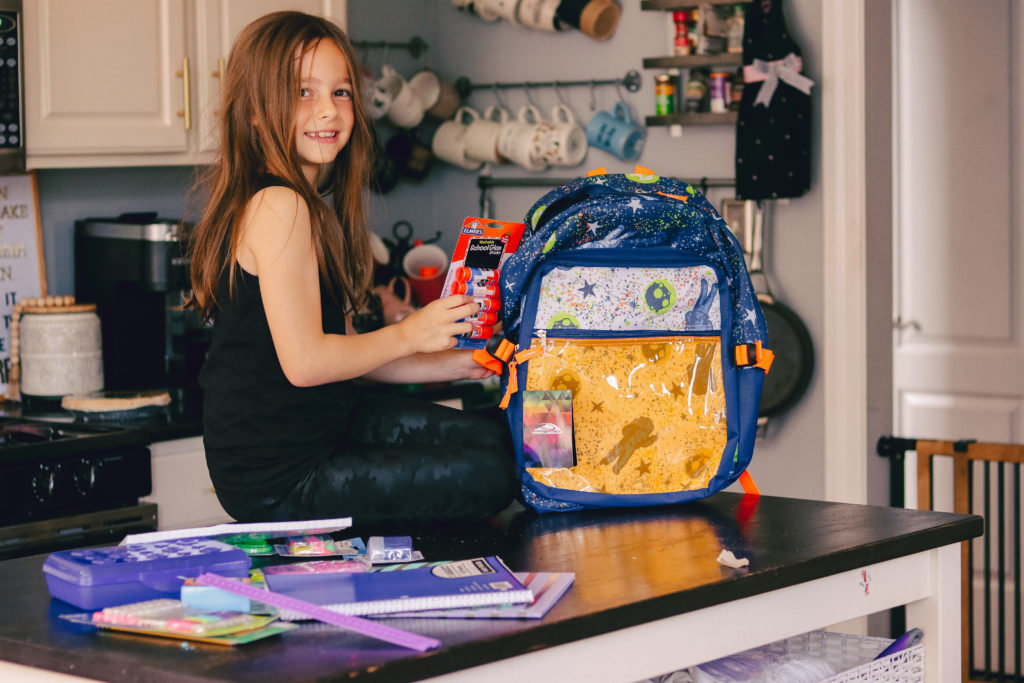 Back to School Tips by popular Nashville lifestyle blog, Nashville Wifestyles: image of a girl sitting on her kitchen counter and putting Dollar General school supplies in her Dollar General backpack. 