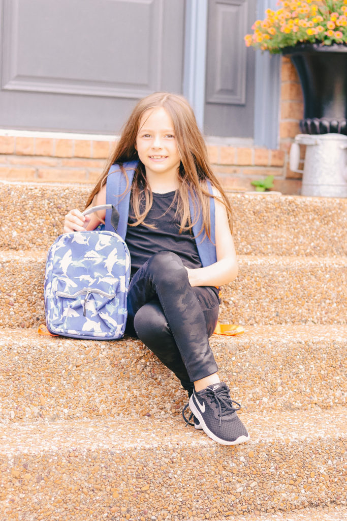Back to School Tips by popular Nashville lifestyle blog, Nashville Wifestyles: image of a little girl wearing a Dollar General backpack and holding a Dollar General lunch box while sitting on her front steps. 