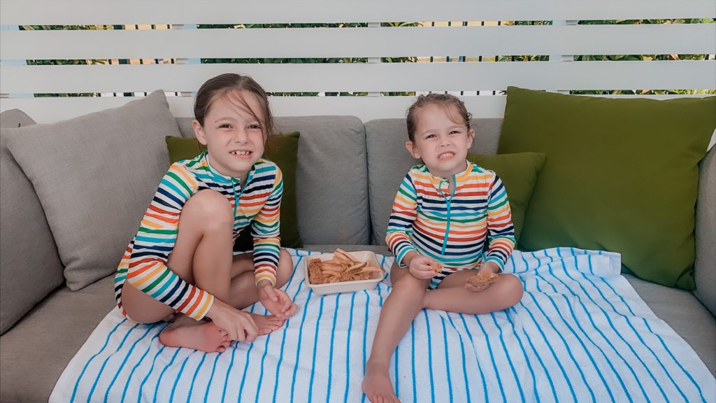 Soundwaves Water Park by popular Nashville blog, Nashville Wifestyles: image of two young sisters wearing matching rainbow strip rash guards and sitting on a blue and white stripe towel while they eat some chicken fingers and french fries. 