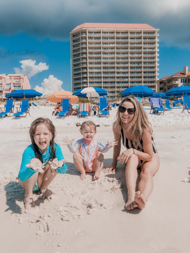 Sun Tanning Tips by popular Nashville beauty blog, Nashville Wifestyles: image of a woman and her daughters wearing their swimsuits and playing in the sand in front of some blue shad umbrellas and blue lounge chairs. 