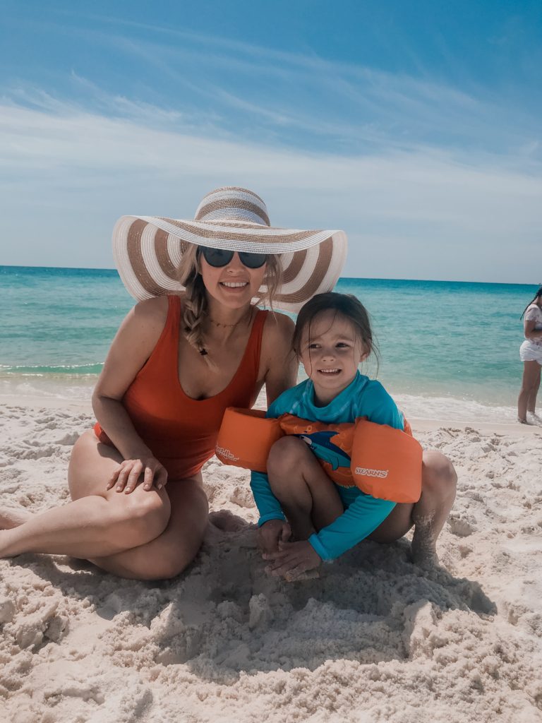 Travel Tips by popular Nashville travel blog, Nashville Wifestyles: image of a woman wearing a red swimsuit and a brown and white stripe sun hat while sitting next to her daughter on the beach. 