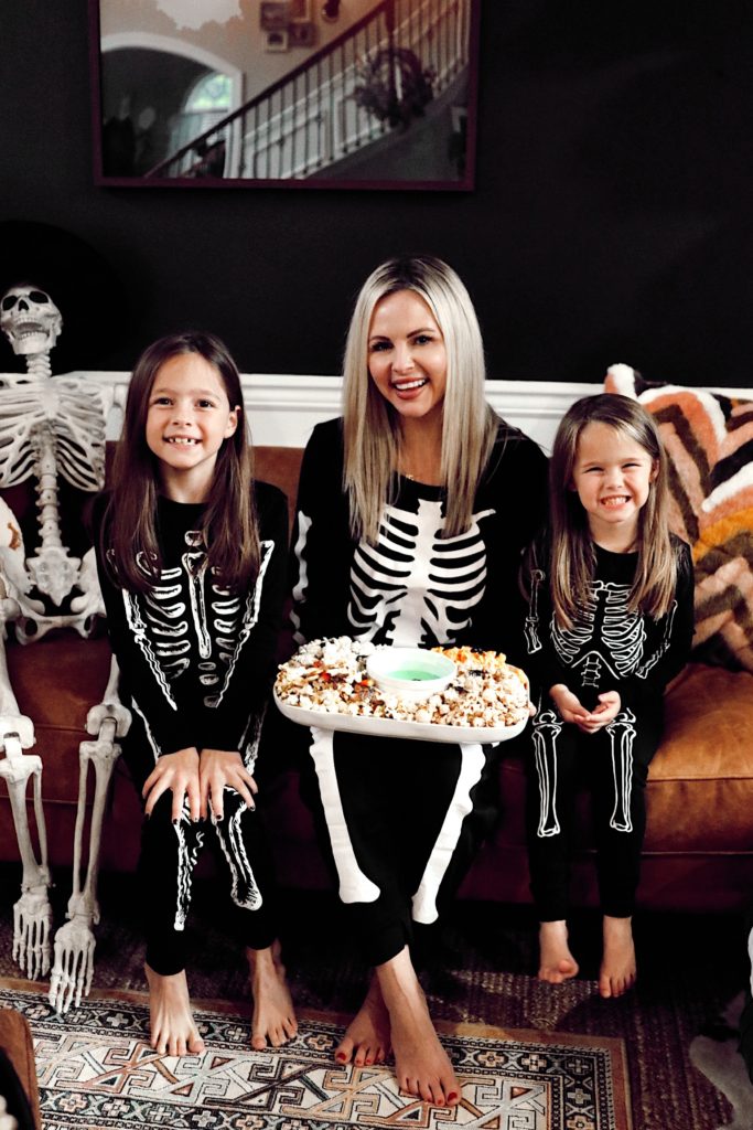 Halloween Pajamas by popular Nashville fashion blog, Nashville Wifestyles: image of a mom and her two daughters wearing skeleton pajamas and sitting on their couch and holding a platter filled with popcorn.  