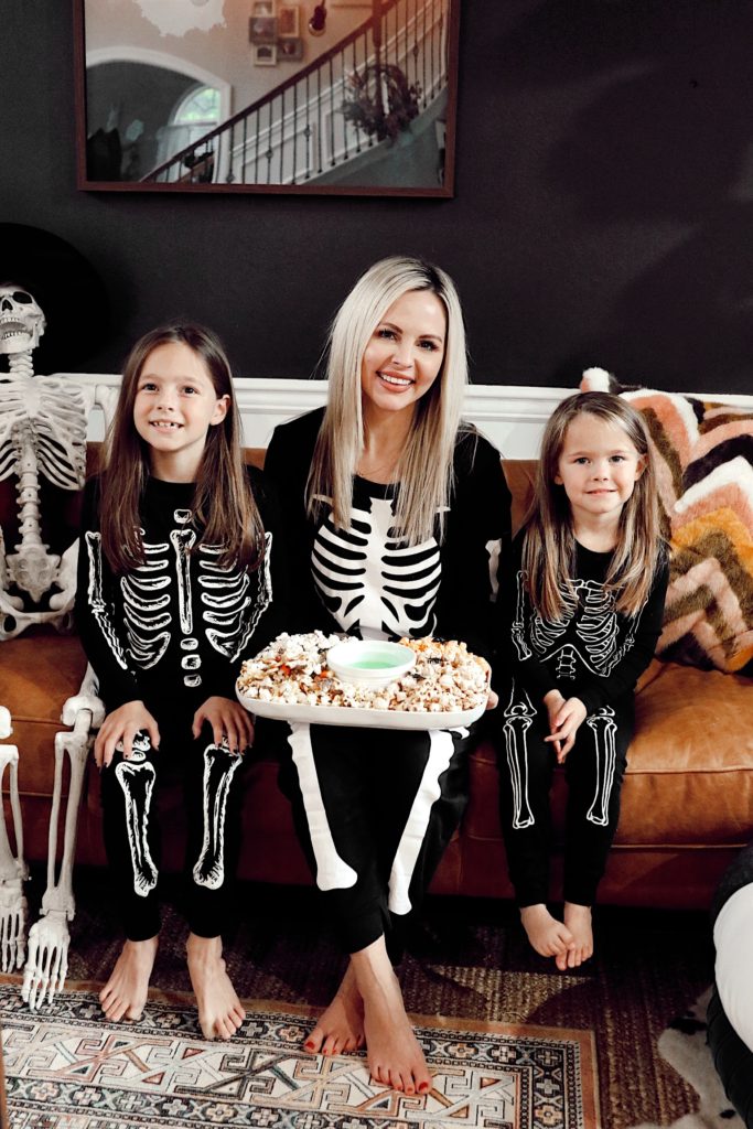 Halloween Pajamas by popular Nashville fashion blog, Nashville Wifestyles: image of a mom and her two daughters wearing skeleton pajamas and sitting on their couch and holding a platter filled with popcorn.  