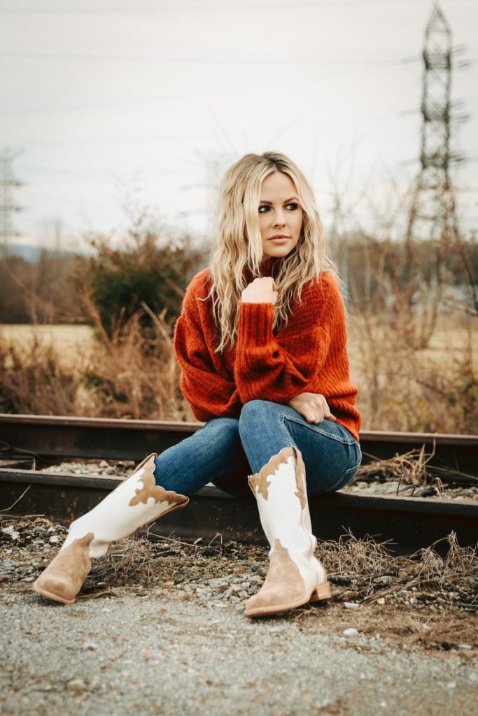 Cowboy Boots for Women by popular Nashville fashion blog, Nashville Wifestyles: image of a woman sitting on a railroad track and wearing a burnt orange sweater, jeans, and tan and white cowboy boots. 