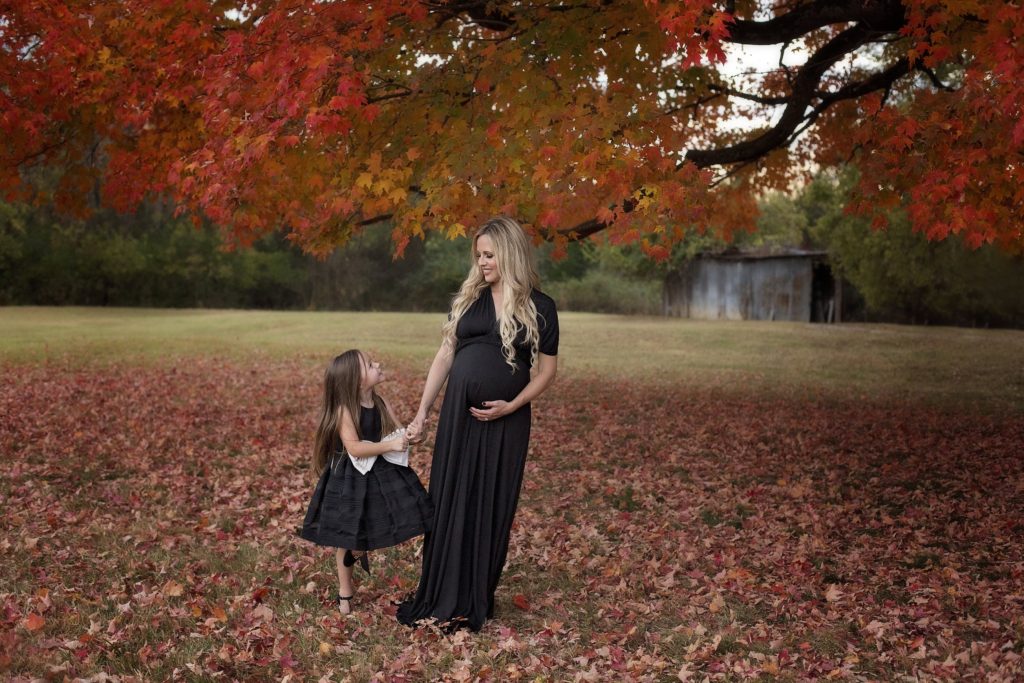Family Maternity Photo Shoot Ideas by popular Nashville lifestyle blog, Nashville Wifestyles: image of pregnant woman wearing a black max dress and a little girl wearing a black sleeveless dress with a large white bow standing together under a tree with red and orange leaves. 