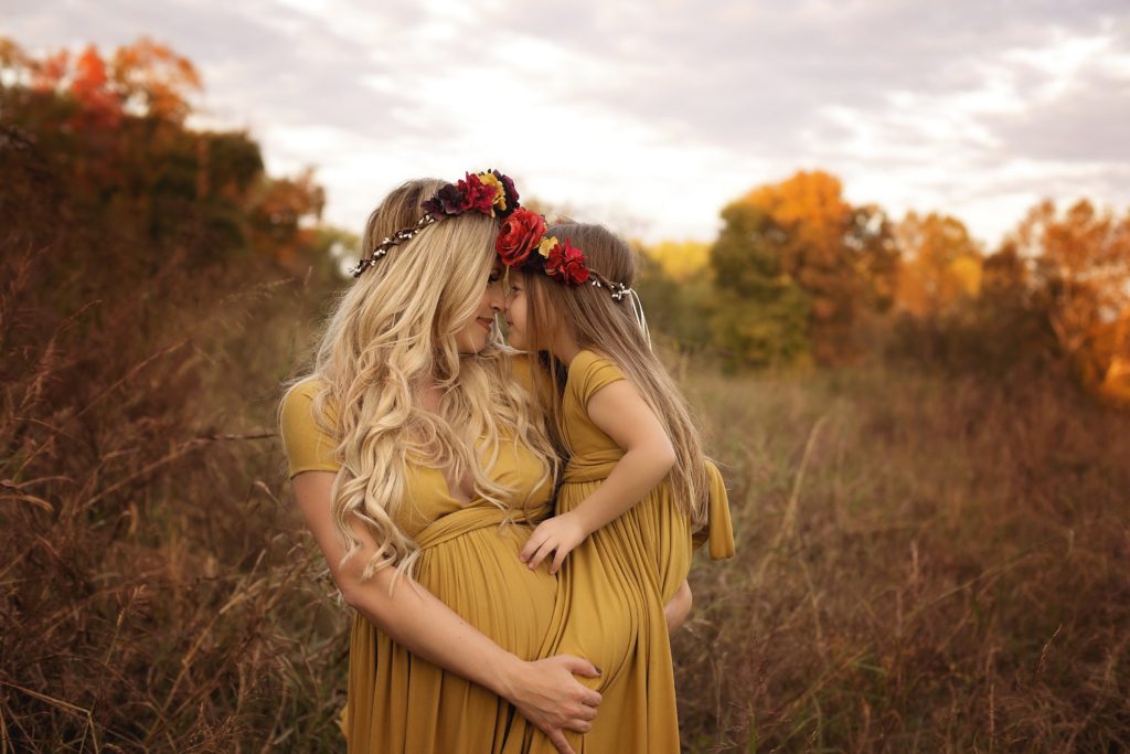 Family Maternity Photo Shoot Ideas by popular Nashville lifestyle blog, Nashville Wifestyles: image of a pregnant woman holding her daughter in a field and both of them wearing matching gold colored maxi dresses and floral crowns. 