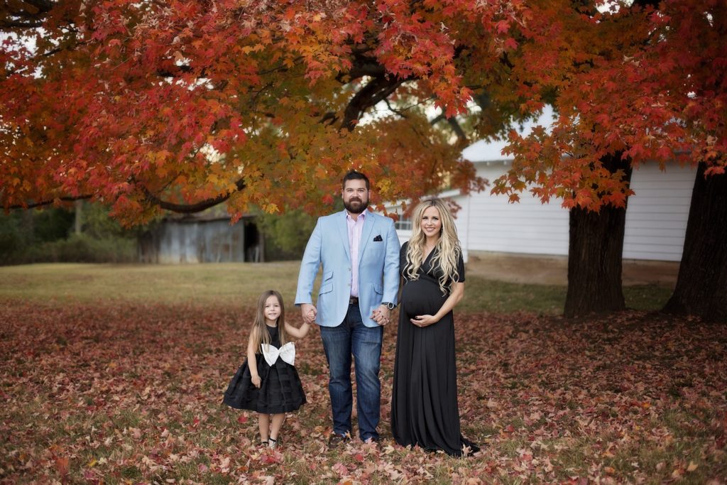 Family Maternity Photo Shoot Ideas by popular Nashville lifestyle blog, Nashville Wifestyles: image of pregnant woman wearing a black max dress, a man wearing a light blue blazer and jeans, and a little girl wearing a black sleeveless dress with a large white bow standing together under a tree with red and orange leaves. 