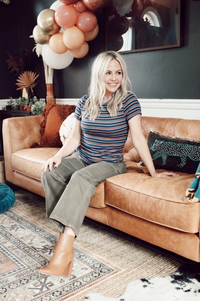 Hoffmann Brothers Home Protection Plan by popular Nashville lifestyle blog, Nashville Wifestyles: image of a woman sitting on her camel brown leather couch and wearing a multi color stripe shirt, olive green pants, and brown ankle boots. 