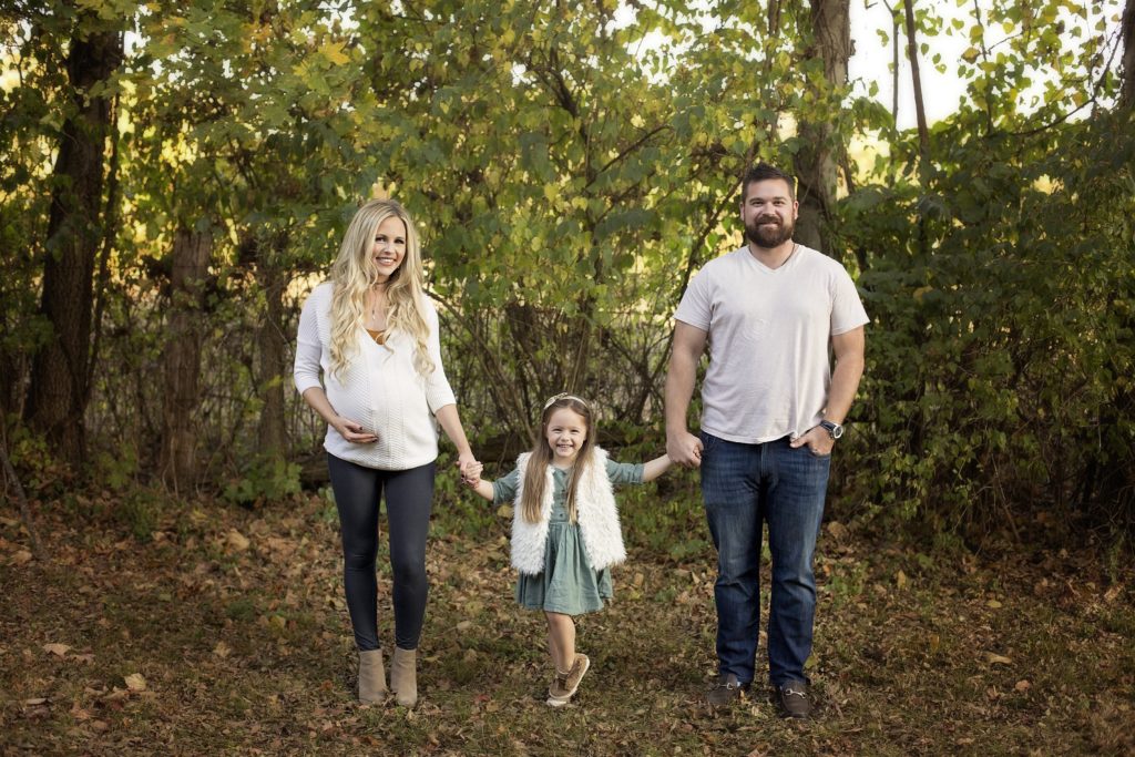 Family Maternity Photo Shoot Ideas by popular Nashville lifestyle blog, Nashville Wifestyles: image of a pregnant woman and her husband holding their young daughter's hands as they stand in a forest. 