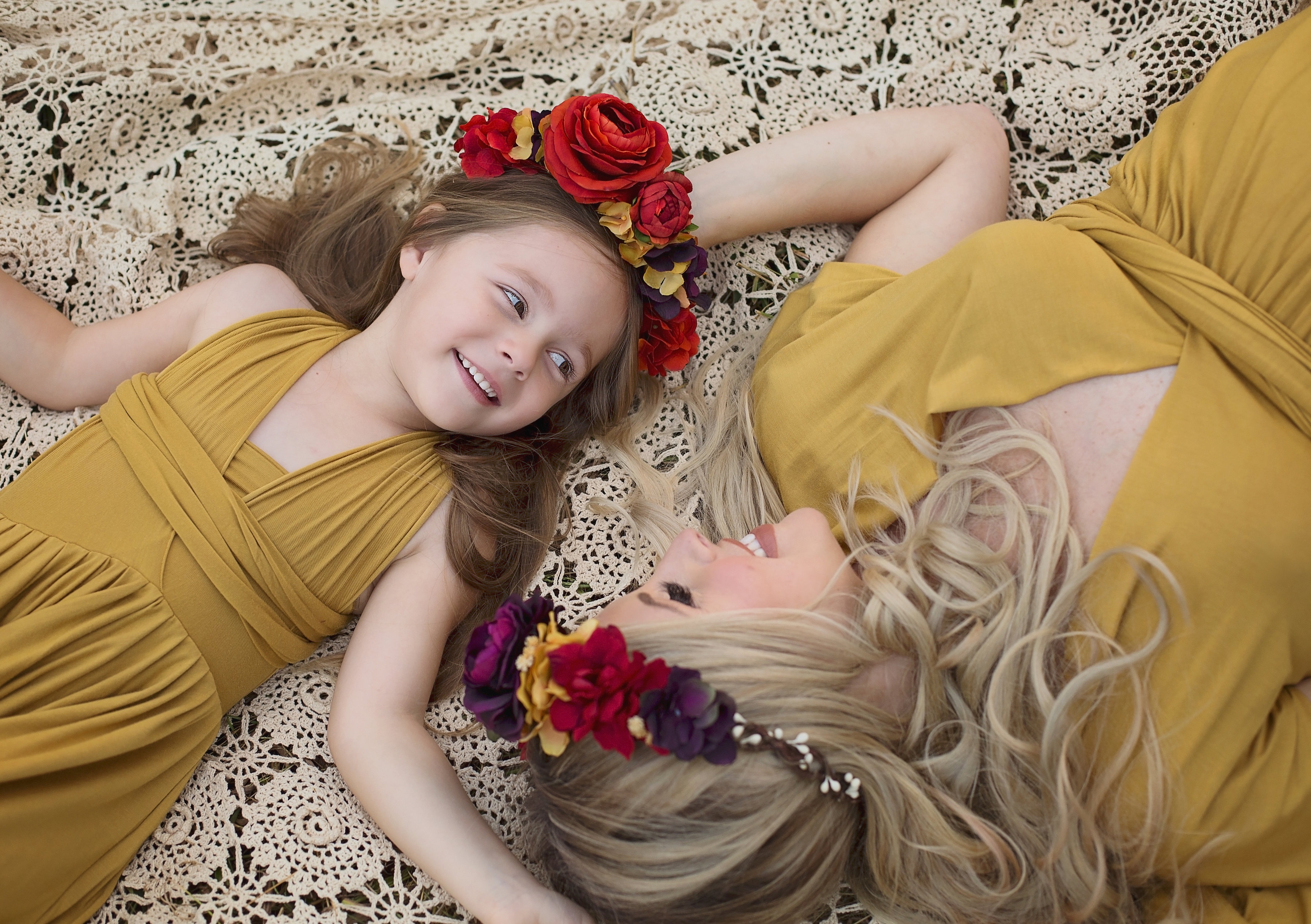 Family Maternity Photo Shoot Ideas by popular Nashville lifestyle blog, Nashville Wifestyles: image of a pregnant woman laying on a lace blanket with daughter in a field and while both of them wear matching gold colored maxi dresses and floral crowns. 