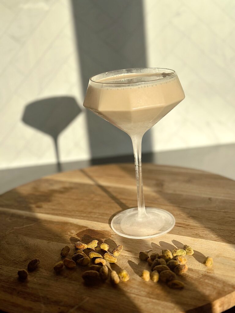 Pistachio Espresso Martini topped with crushed pistachios in a chilled martini glass, creamy coffee cocktail on marble surface with coffee beans and gold shaker nearby